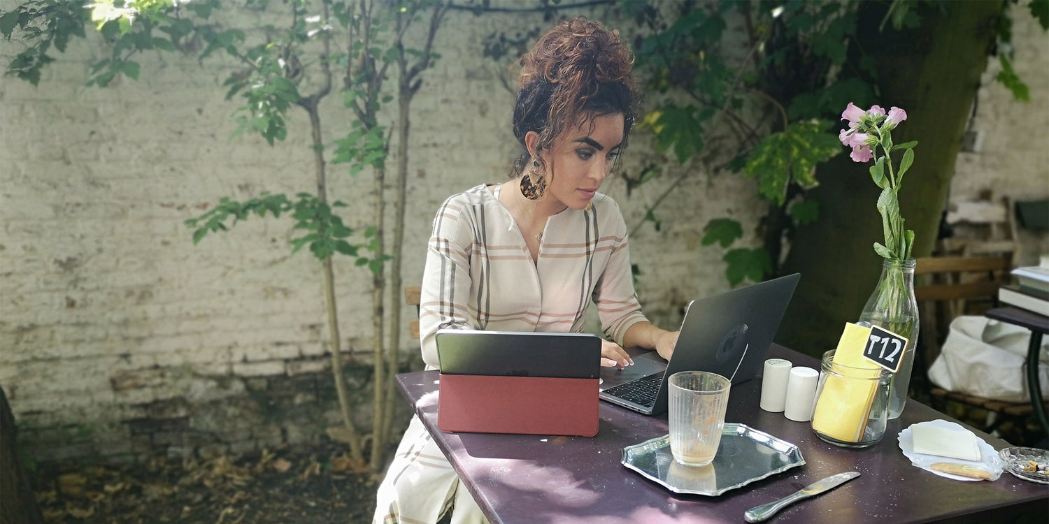 Woman working outdoors at a café table with a laptop and tablet, surrounded by greenery and a vase with pink flowers.