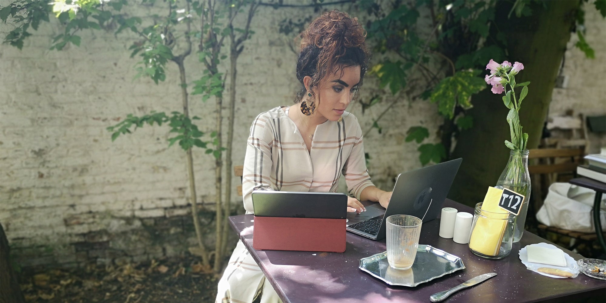 Woman working outdoors at a café table with a laptop and tablet, surrounded by greenery and a vase with pink flowers.