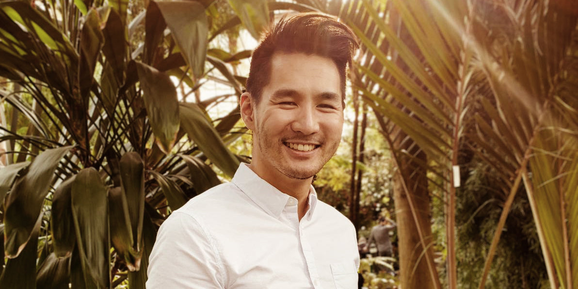 Smiling man in a white button-up shirt standing outdoors surrounded by tropical plants, sunlight filtering through the leaves.