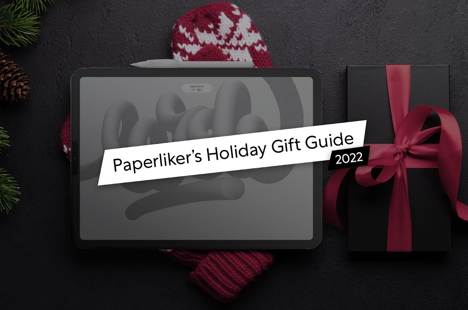 iPad with Apple Pencil resting on top, placed on holiday-themed background with pine branches, pinecones, red knit accessories, and a wrapped gift box with a red ribbon. Text overlay reads ‘Paperliker’s Holiday Gift Guide 2022.’