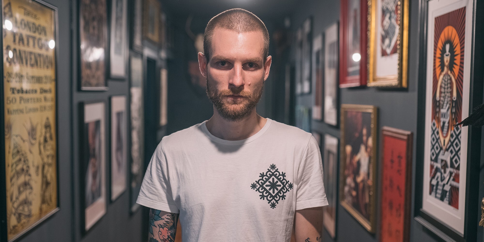 Serious-looking man with a short beard and buzzcut wearing a white t-shirt with a black geometric design, standing in a hallway lined with framed tattoo artwork.