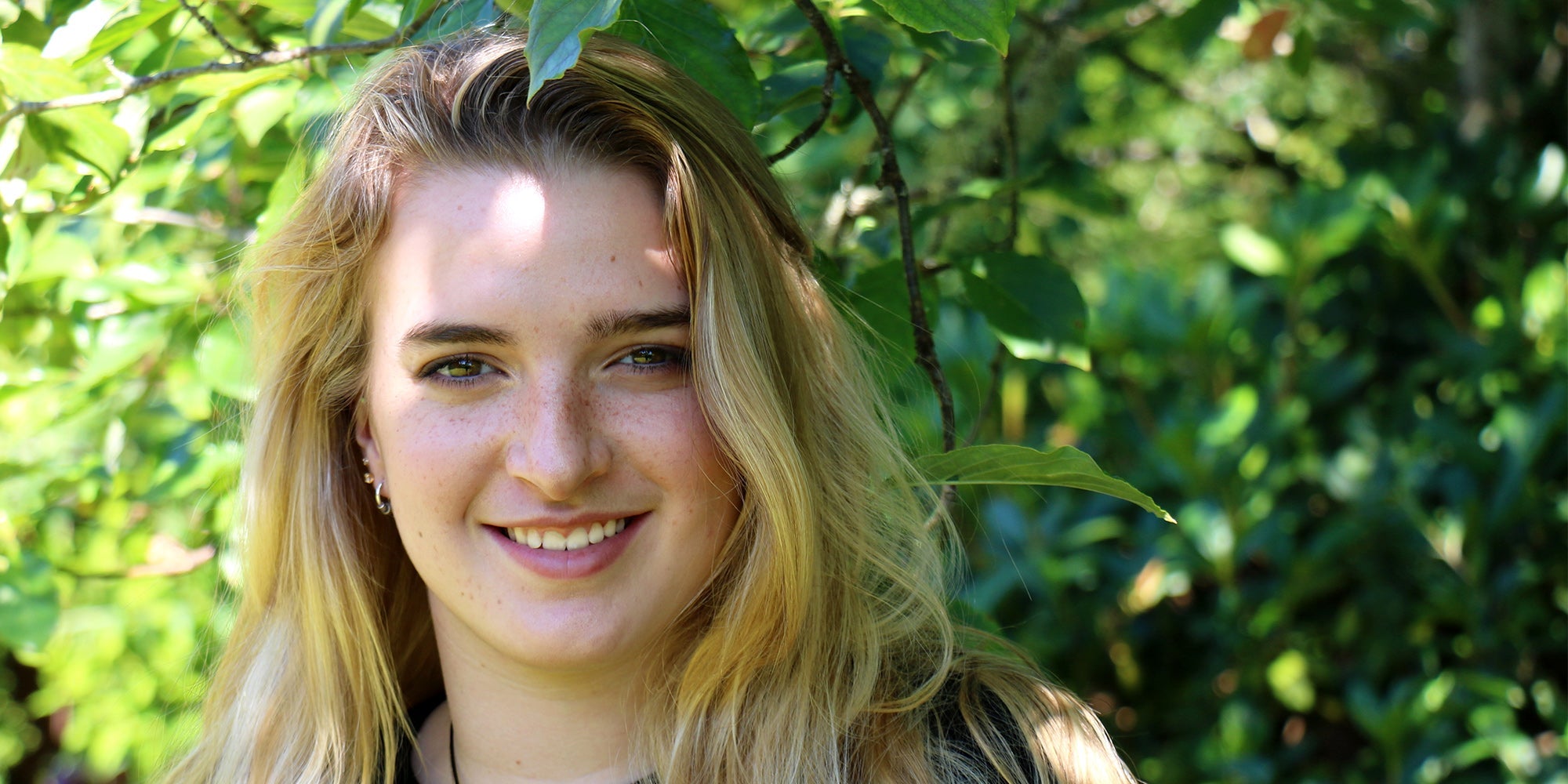 Portrait of a young woman smiling outdoors with sunlight and green leaves in the background.