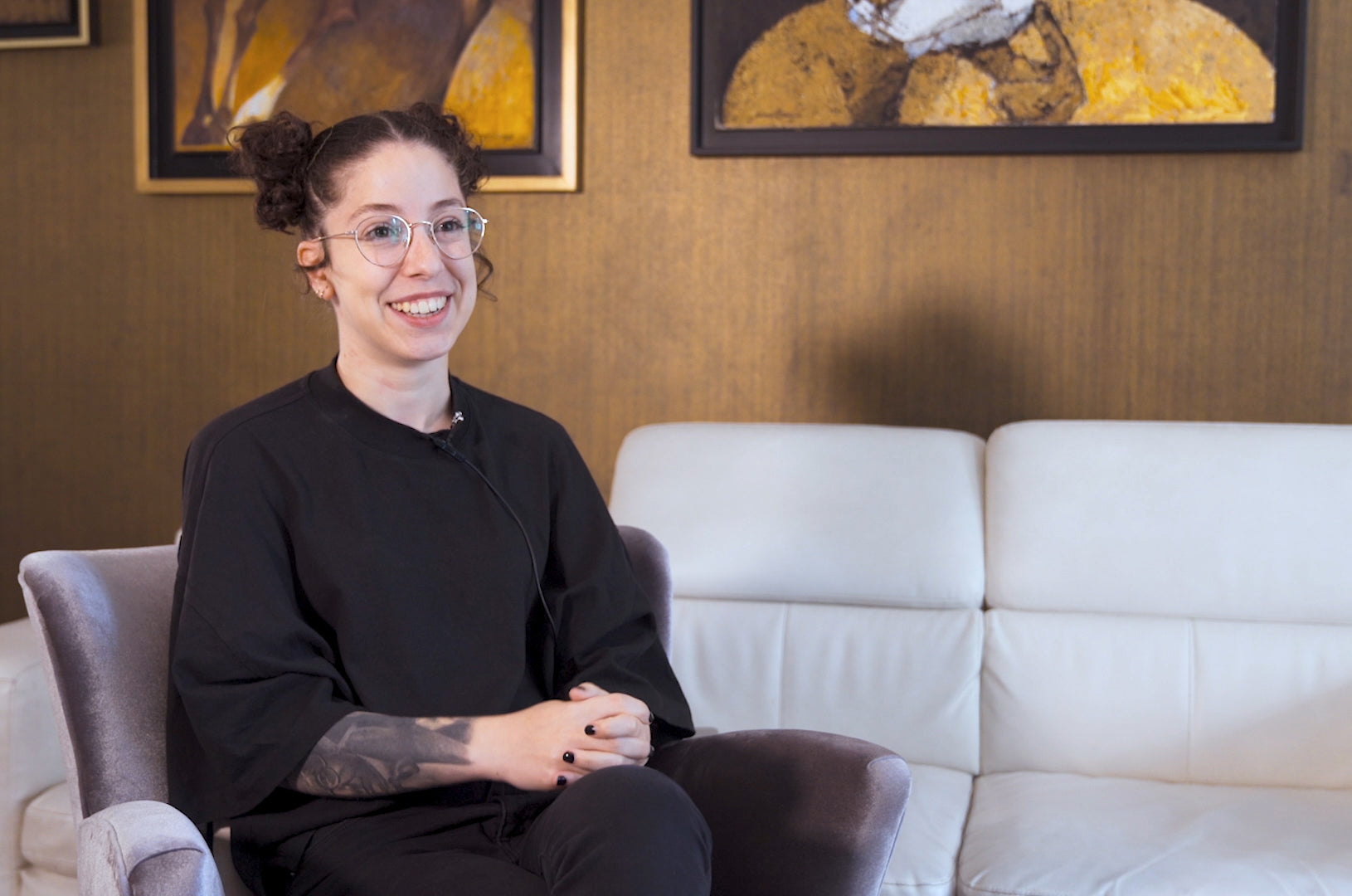 Smiling person with glasses and tattoos, seated indoors on a chair with artwork on the wall behind them.