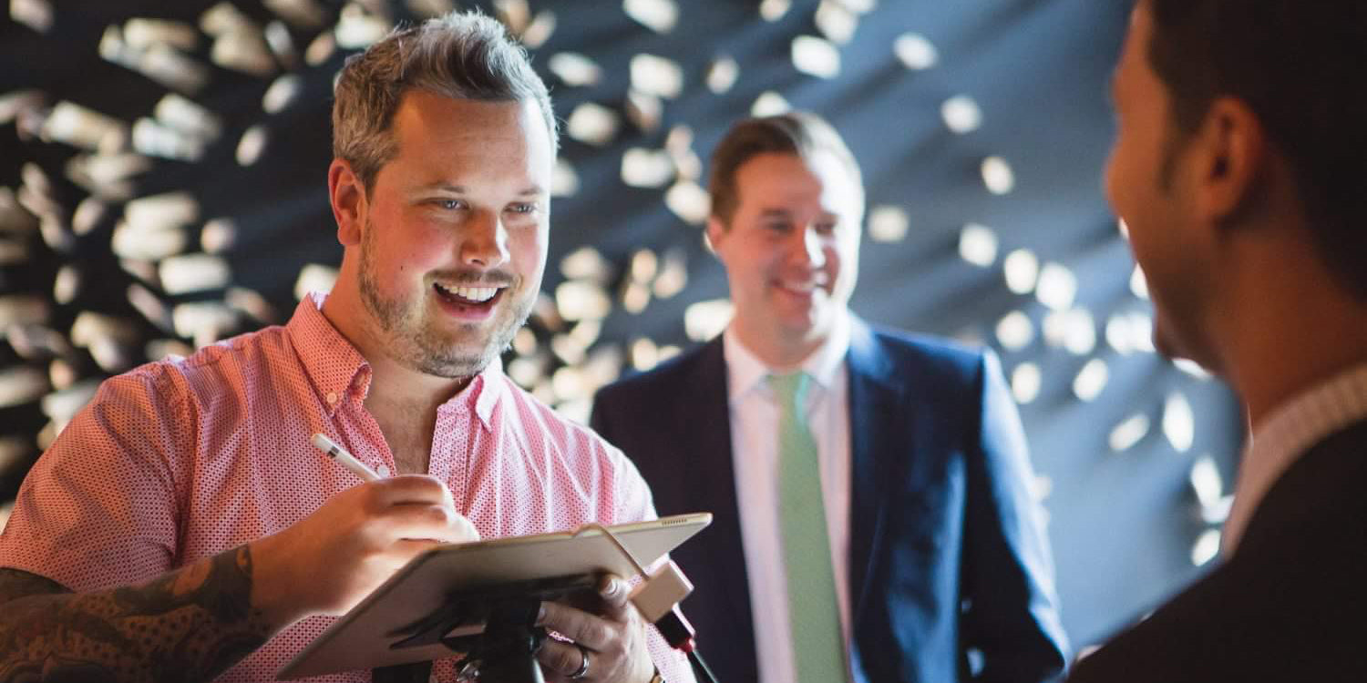 Man smiling while drawing on an iPad with Apple Pencil at an event, with two other men in the background.