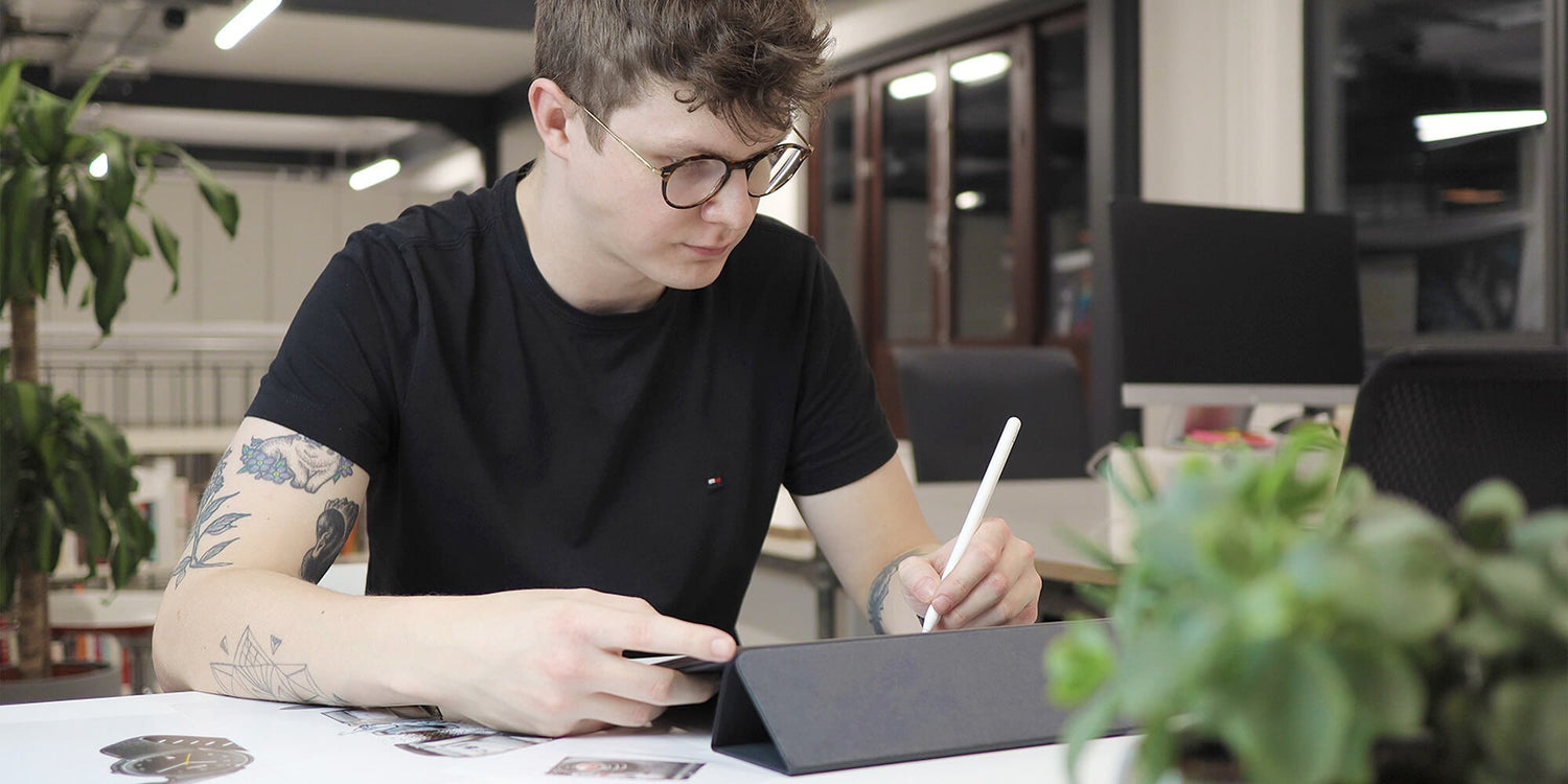 Man with glasses and tattoos drawing on an iPad with an Apple Pencil at a desk in an office setting.