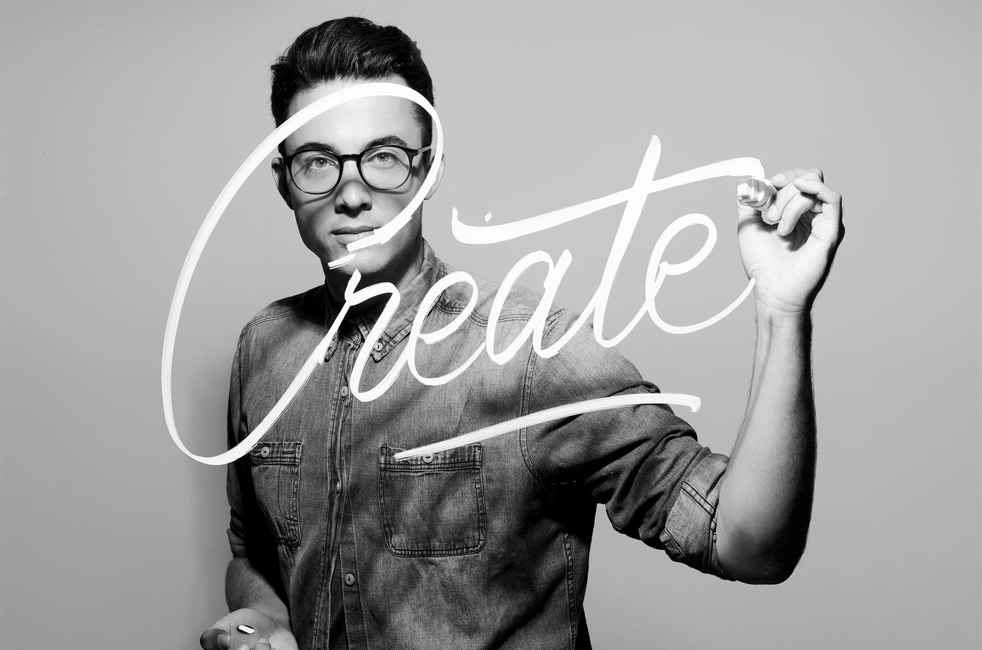Black and white photo of a man in glasses writing the word ‘Create’ in large script across the image.