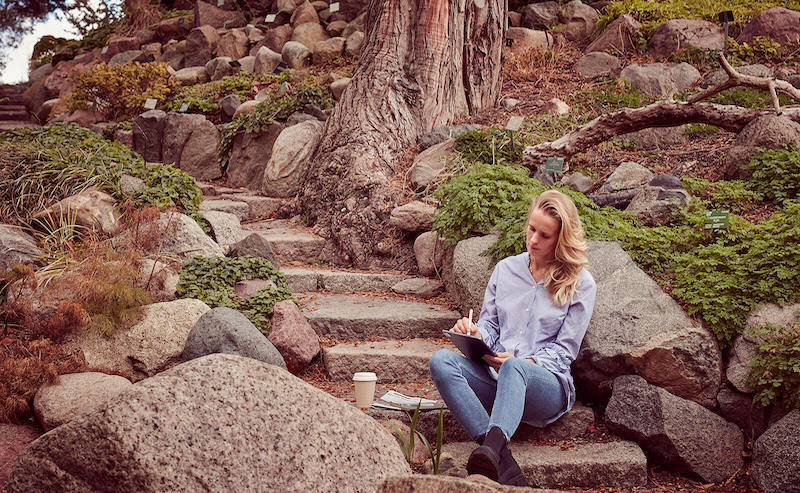 A woman sits outside on concrete steps in a scenic outdoor environment while writing on an iPad with an Apple Pencil.
